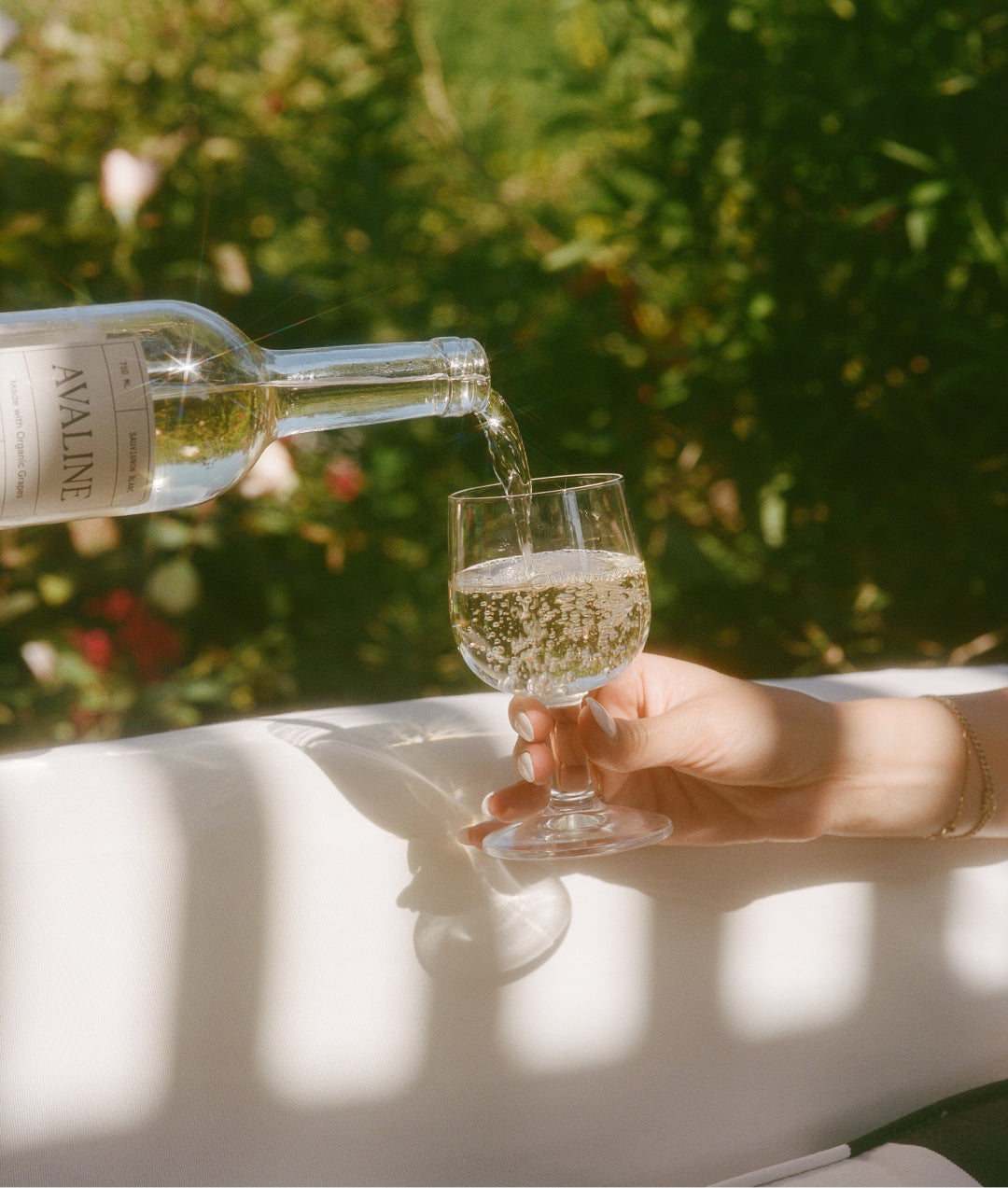 An image of a bottle of Sauvignon Blanc being poured into a wine glass held by a woman’s hand, with greenery and flowers in the background.