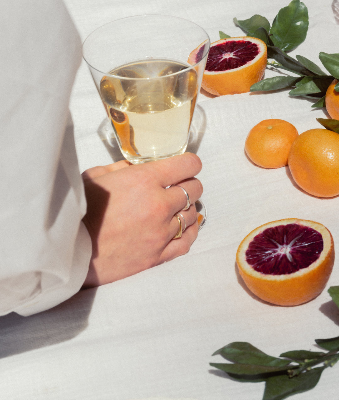 An image of a woman's hand holding a glass of white wine outside on a blanket with oranges and greenery